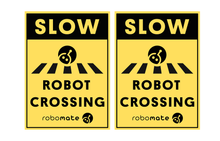 wo yellow Robot Crossing signs with black robot mower icons and mounting stakes, positioned beside a driveway to alert drivers to robots crossing